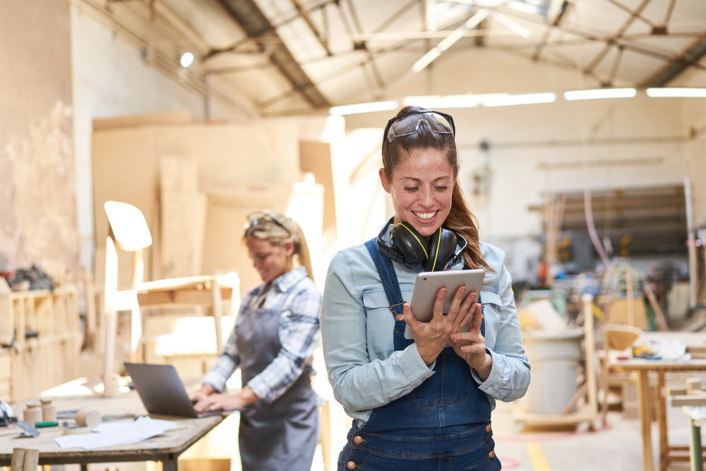 Dwie uśmiechnięte młode kobiety w warsztacie stolarskim wykorzystują urządzenia mobilne do pracy